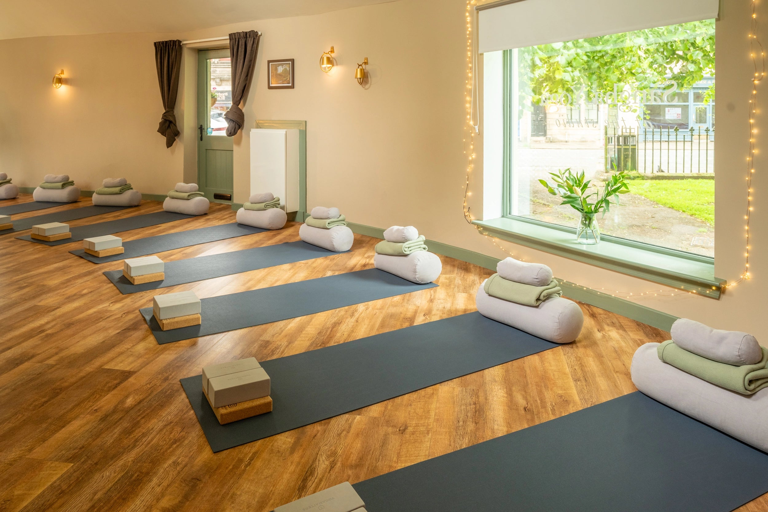 A yoga studio with multiple dark blue yoga mats arranged in rows on a wooden floor, each with folded white and green towels and a block. A large window shows greenery outside, and string lights decorate the wall.