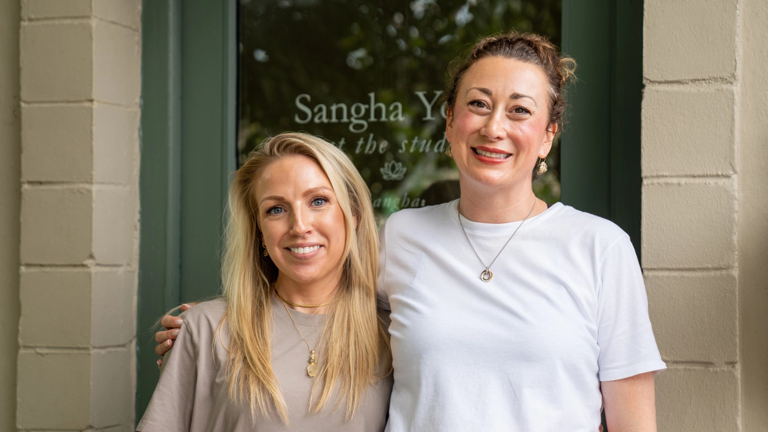 Two women smiling and posing together in front of a storefront with a dark green door and beige brick columns. The woman on the right wears a plain white t-shirt and necklace, while the woman on the left wears a taupe-beige t-shirt and gold pendant necklace.
