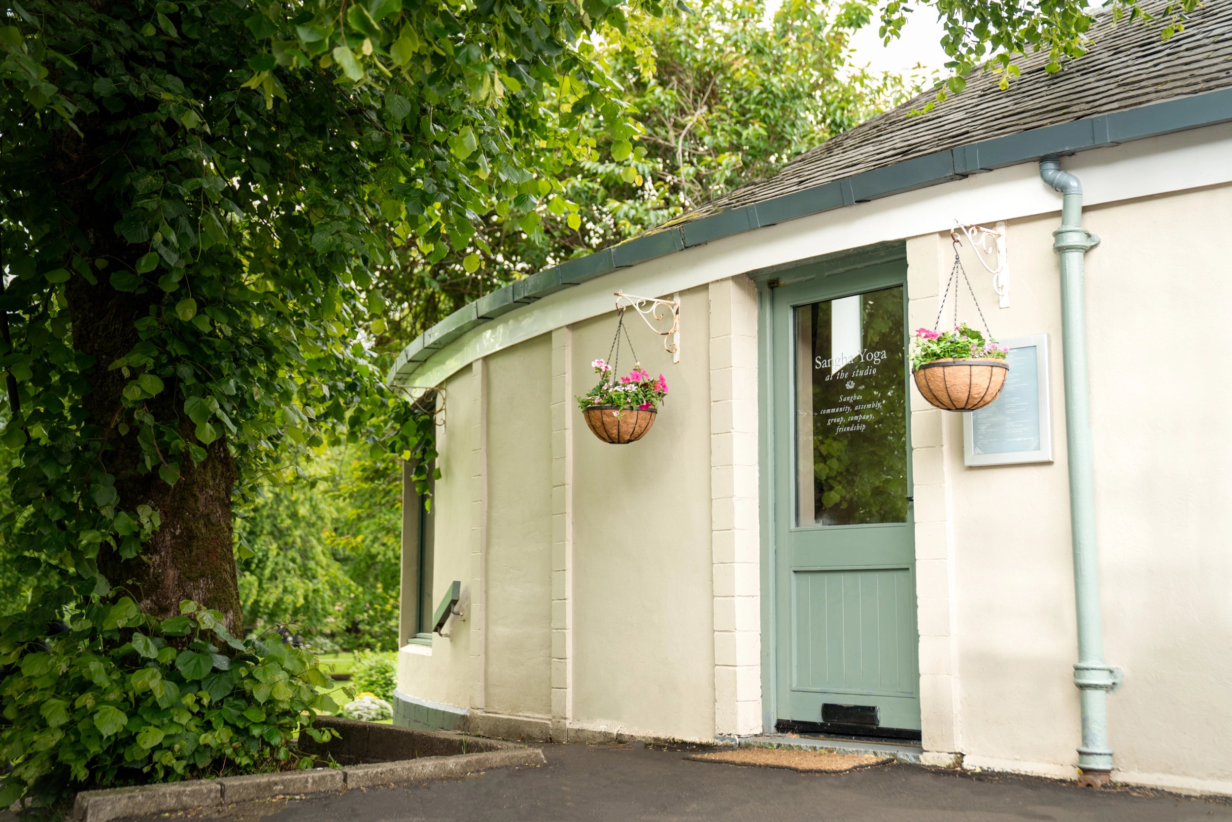 A small cream-colored building with a curved wall, a pale green door, hanging flower baskets, and dense green foliage around it.