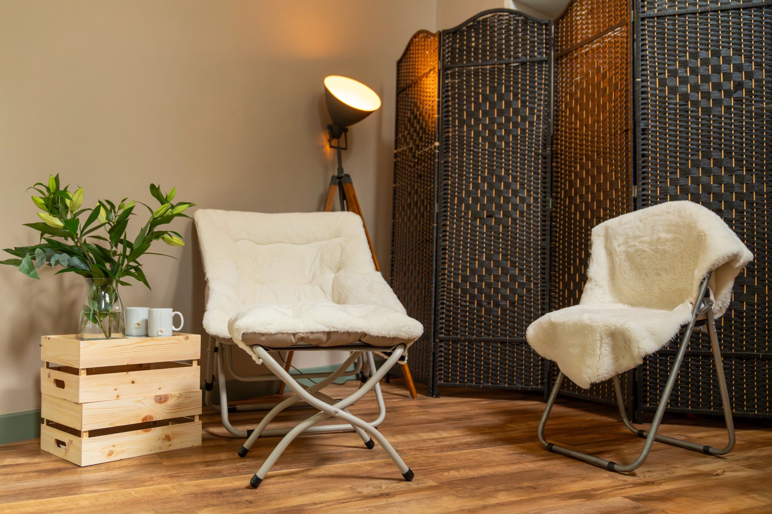Two modern folding camp chairs with cream sheepskin-style cushions and silver metal frames, styled in a warm-toned room with a wooden floor, a black folding screen, a floor lamp, and a small wooden crate holding a plant and mugs.